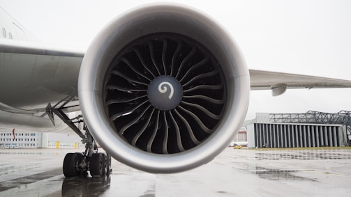 A large airplane engine with turbine blades is prominently displayed, mounted on the wing of an aircraft. The aircraft is stationed on a wet tarmac near an airport hangar. The detailed view highlights the intricate design of the engine blades, and in the background, parts of the wing and wheels of the aircraft are visible.