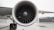 A large airplane engine with turbine blades is prominently displayed, mounted on the wing of an aircraft. The aircraft is stationed on a wet tarmac near an airport hangar. The detailed view highlights the intricate design of the engine blades, and in the background, parts of the wing and wheels of the aircraft are visible.