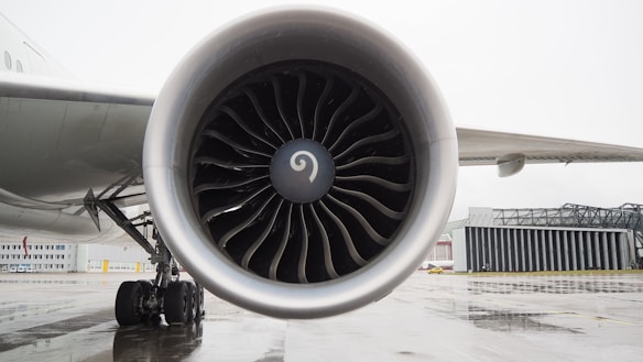A large airplane engine with turbine blades is prominently displayed, mounted on the wing of an aircraft. The aircraft is stationed on a wet tarmac near an airport hangar. The detailed view highlights the intricate design of the engine blades, and in the background, parts of the wing and wheels of the aircraft are visible.