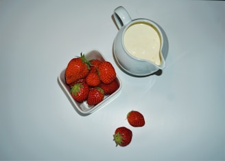 A white pitcher filled with cream or milk sits beside a square white bowl containing several ripe red strawberries. Two additional strawberries are placed on the surface in front of the pitcher and bowl.