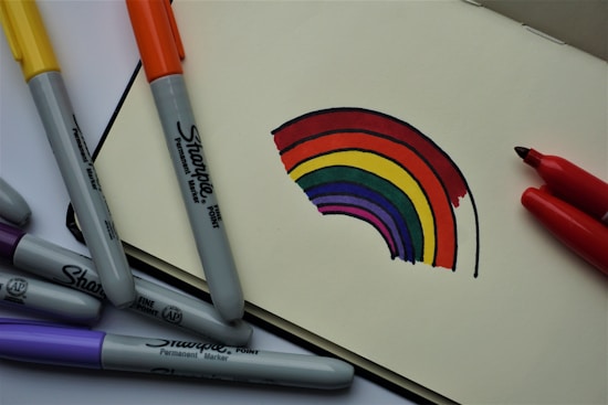 A partially drawn rainbow with vibrant colors is on a sketchbook page. Surrounding the sketchbook are several Sharpie permanent markers in various colors, including yellow, orange, purple, and red. The markers are positioned casually around the drawing, suggesting an ongoing art project.