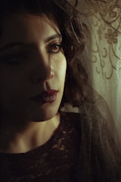 A thoughtful woman gazes into the distance, with soft lighting creating shadows on her face. Her eyes are accented with dark makeup, and her lips are painted in a deep shade. The background features a lace curtain with floral patterns, adding a sense of vintage elegance.