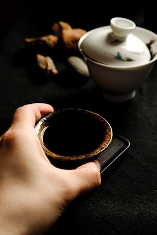 A hand holding a small, rustic-looking ceramic cup filled with a dark liquid. Beside it, there is a porcelain cup with a lid and minimal decoration. The background appears to be dark, with what seems to be pieces of wood or stones placed subtly in a blurred fashion.