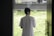 A person wearing a light-colored shirt stands inside, facing a window with a view of grass and a parked car. The lighting suggests a calm and contemplative atmosphere.