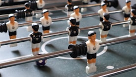 A close-up view of a foosball table with several plastic soccer player figures mounted on rods. The figures are dressed in black and white uniforms, positioned on a green surface resembling a soccer field. A small blue and white soccer ball is visible among the figures.