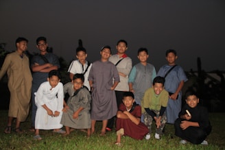 A group of boys is standing and kneeling on a grassy field, wearing various traditional outfits. It is nighttime or dusk, with a dark sky in the background, and the scene is slightly illuminated by an artificial light source. The boys are smiling and appear to be posing for the photo.