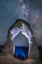 A night sky featuring the Milky Way galaxy stretches across a clear, star-filled backdrop. The scene is dominated by a large, naturally occurring stone arch formation illuminated by two people standing at its base with bright blue lights. The cosmic and terrestrial elements create a sense of wonder and scale.