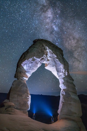 A night sky featuring the Milky Way galaxy stretches across a clear, star-filled backdrop. The scene is dominated by a large, naturally occurring stone arch formation illuminated by two people standing at its base with bright blue lights. The cosmic and terrestrial elements create a sense of wonder and scale.