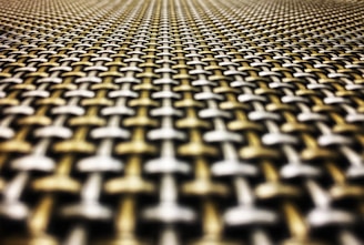 A close-up perspective of a textured woven pattern made of interlocking metal strands creating a geometric and symmetrical design. The image emphasizes the depth and detail of the material, with a gradient blur making it appear to stretch into the distance.