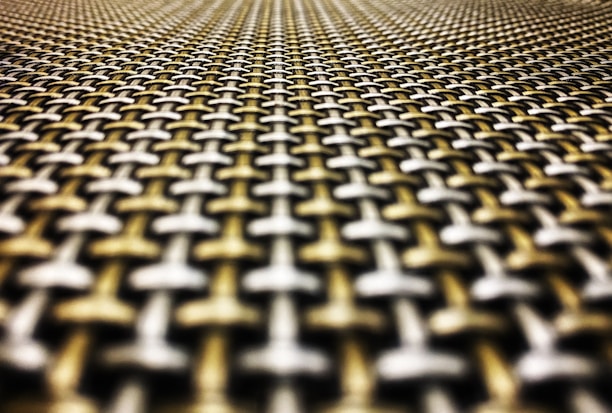 A close-up perspective of a textured woven pattern made of interlocking metal strands creating a geometric and symmetrical design. The image emphasizes the depth and detail of the material, with a gradient blur making it appear to stretch into the distance.
