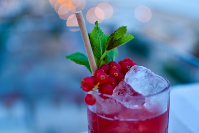 A refreshing cocktail in a glass garnished with a slice of ice, red currants, and fresh mint leaves. The background is blurred, adding a bokeh effect with circular lights.