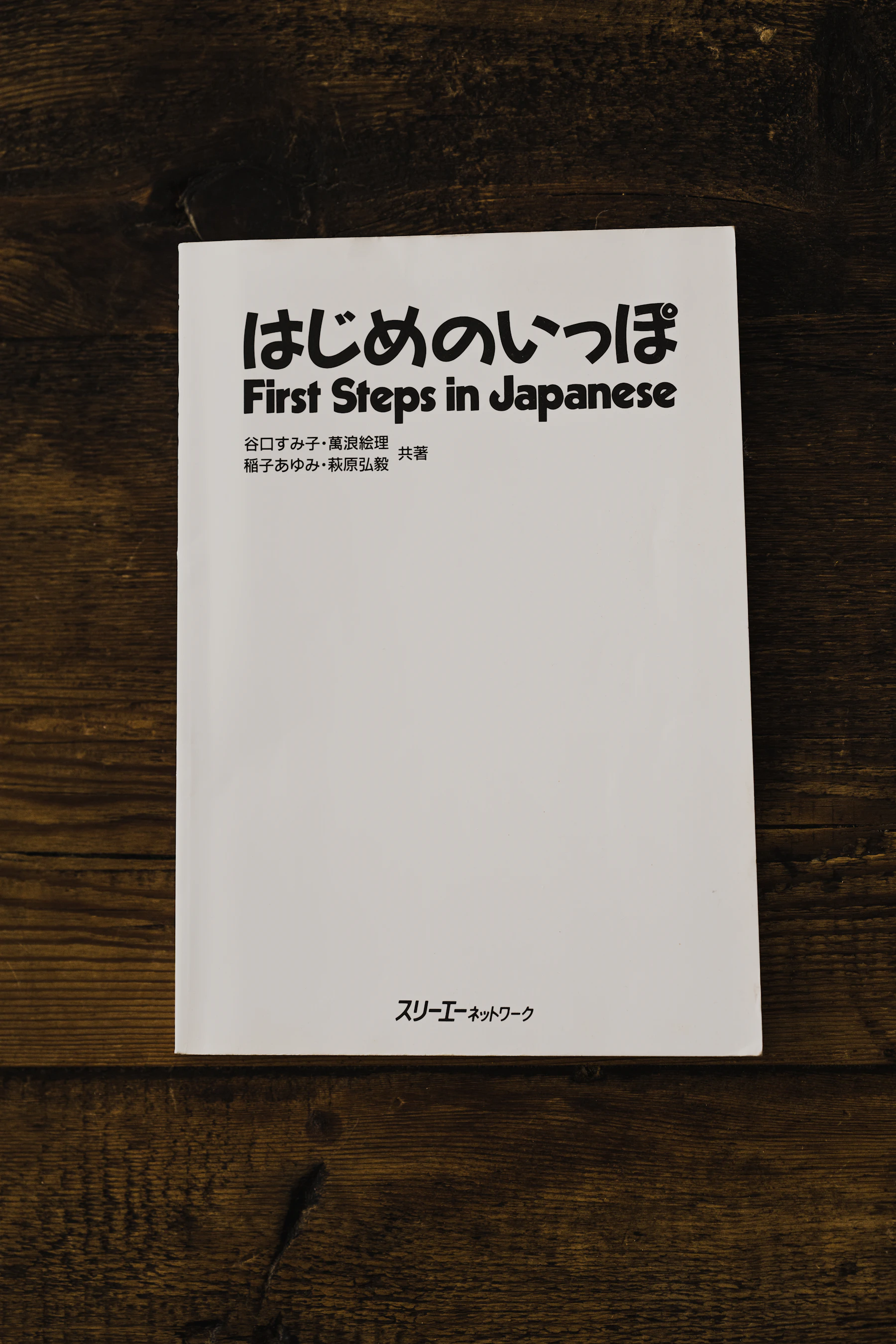 A white booklet titled First Steps in Japanese, in hiragana and in English, lying on a dark wooden table