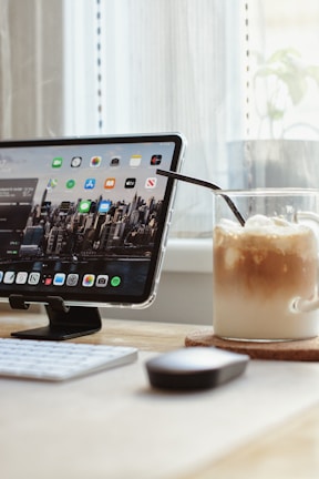 A digital tablet displaying various app icons in a holder on a desk next to a cup of iced coffee with whipped cream and a straw. A wireless keyboard and mouse are visible in the foreground, with a background of a light curtain and a potted plant near the window.