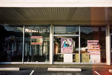 A storefront for a dry cleaning business with large glass windows displaying various signs and clothing racks with garments hung inside. The signage includes 'Nashville's Shirts Laundered' and services like spotting, drapery, silk and wool care. The central window features a colorful poster of a man and woman labeled 'Dry Cleaners.' An American flag is visible in the reflection on the glass.