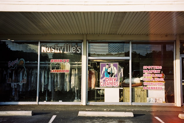A storefront for a dry cleaning business with large glass windows displaying various signs and clothing racks with garments hung inside. The signage includes 'Nashville's Shirts Laundered' and services like spotting, drapery, silk and wool care. The central window features a colorful poster of a man and woman labeled 'Dry Cleaners.' An American flag is visible in the reflection on the glass.