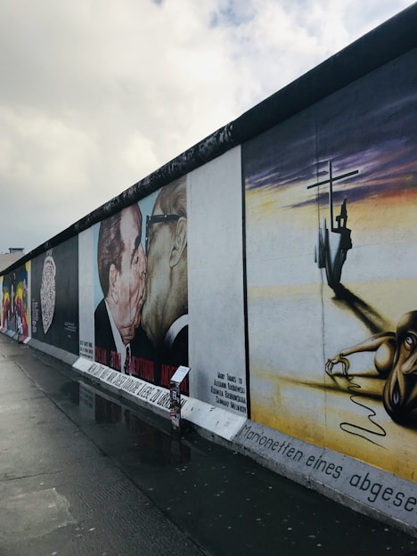 A series of murals painted on a section of a wall, depicting various artistic and political themes. Prominently, one mural shows two men kissing, with text beneath it. Another mural displays a puppet-like figure manipulated by large hands, set against a yellow and white background. The ground is wet, reflecting parts of the murals.