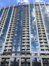 A modern high-rise building with numerous floors and balconies, featuring a sleek design with large glass windows that reflect the blue sky and clouds. The structure has a mix of white and brown paneling, with a sign on top that reads 'DIM'.