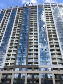 A modern high-rise building with numerous floors and balconies, featuring a sleek design with large glass windows that reflect the blue sky and clouds. The structure has a mix of white and brown paneling, with a sign on top that reads 'DIM'.