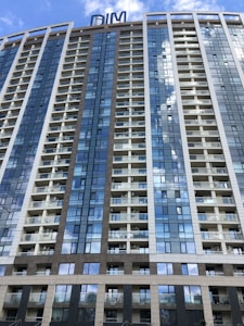 A modern high-rise building with numerous floors and balconies, featuring a sleek design with large glass windows that reflect the blue sky and clouds. The structure has a mix of white and brown paneling, with a sign on top that reads 'DIM'.