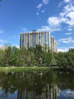A tall, modern building with the word 'DIM' on top is in the background, surrounded by lush green trees. In the foreground, a tranquil body of water reflects the clear blue sky and parts of the building. People are walking along a path near the trees.