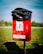 A red outdoor waste bin situated on a grassy area, with a black plastic bag lining the top. The bin features a sign illustrating a person disposing of waste alongside a dog, indicating it is for dog waste. The background is slightly blurred, showing green trees and a blue sky.