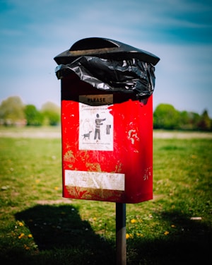 A red outdoor waste bin situated on a grassy area, with a black plastic bag lining the top. The bin features a sign illustrating a person disposing of waste alongside a dog, indicating it is for dog waste. The background is slightly blurred, showing green trees and a blue sky.