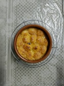 A round ceramic dish filled with a baked, golden-brown dish, possibly featuring individual servings or dumplings. The dish sits on a wire rack atop a textured and patterned tablecloth.