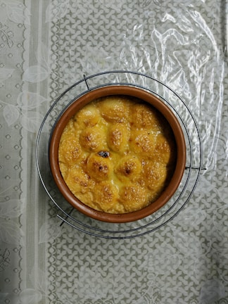 A round ceramic dish filled with a baked, golden-brown dish, possibly featuring individual servings or dumplings. The dish sits on a wire rack atop a textured and patterned tablecloth.