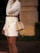 A person wearing a white double-breasted coat dress with a gold belt and holding a beige handbag. The coat dress is complemented by an elegant hairstyle featuring a long braid. The scene appears to be set outdoors, with shrubs and a paved area in the background.