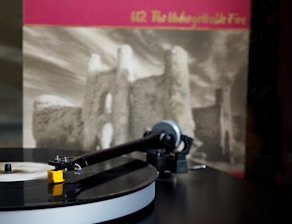 A vinyl record is being played on a turntable with a blurred album cover in the background. The turntable's tonearm and stylus are visible, and the vinyl has a glossy surface. The album cover has an image of ruins and sky, with text at the top.