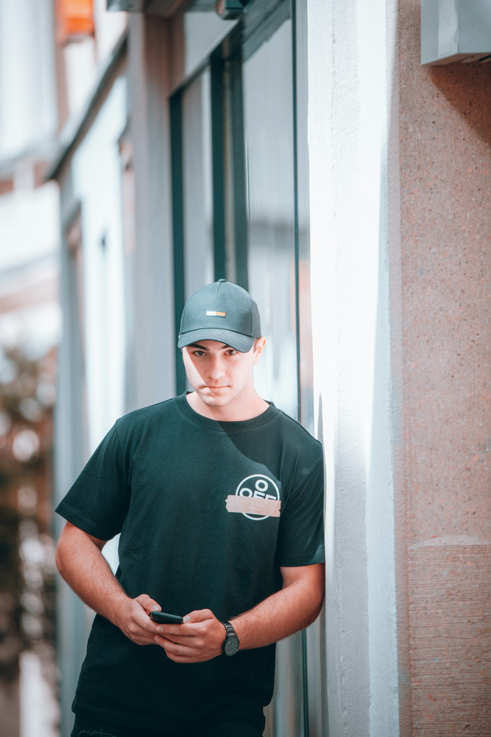 man in black crew neck t-shirt wearing blue cap standing beside white wall during daytime