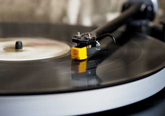 A close-up of a vinyl record on a turntable with a yellow tonearm and a needle gently placed on the grooves of the black record. The background is blurred, focusing attention on the details of the turntable mechanism.