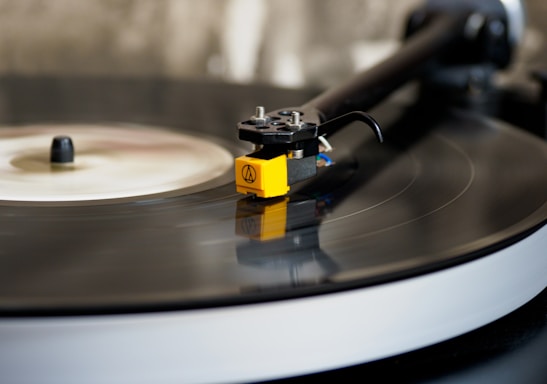 A close-up of a vinyl record on a turntable with a yellow tonearm and a needle gently placed on the grooves of the black record. The background is blurred, focusing attention on the details of the turntable mechanism.