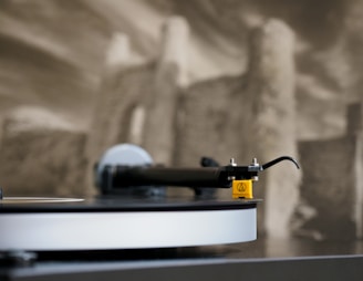 A close-up view of a turntable's tonearm stylus on a vinyl record, with a blurred monochrome background depicting a rustic building or ruin.