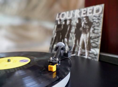 A vinyl record is spinning on a turntable with a yellow label and an Audio-Technica cartridge. In the background, a black and white album cover with the name 'Lou Reed' is visible, showing figures in a slightly blurred style.