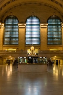 A grand hall with tall arched windows featuring intricate grid-like patterns allows soft natural light to enter. The centerpiece is a circular information booth with a prominent clock on top, surrounded by an elegant beige interior with columns and marble floors. A few people are sparsely present in the background.