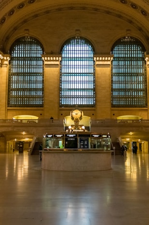 A grand hall with tall arched windows featuring intricate grid-like patterns allows soft natural light to enter. The centerpiece is a circular information booth with a prominent clock on top, surrounded by an elegant beige interior with columns and marble floors. A few people are sparsely present in the background.