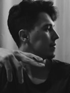 A black and white portrait of a person in profile with short, dark hair. The subject's hand is partially raised near the face, creating a casual, thoughtful pose. The background is softly blurred, emphasizing the person's features.