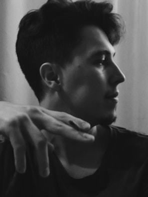 A black and white portrait of a person in profile with short, dark hair. The subject's hand is partially raised near the face, creating a casual, thoughtful pose. The background is softly blurred, emphasizing the person's features.
