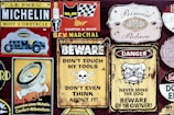 A collection of vintage signs and advertisements, featuring brands like Michelin and SEV Marchal, along with humorous and cautionary messages such as 'Don't Touch My Tools' and 'Beware of the Owner'. The signs are colorful with a weathered look, featuring vintage logos and illustrations, including a cat, a tire person, and a fierce bulldog.