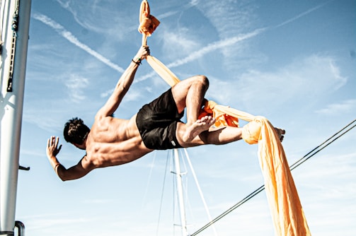 A person is suspended in mid-air using bright orange aerial silks, performing an acrobatic pose against a backdrop of a clear blue sky. The individual appears to be on a sailboat, suggested by the visible mast and rigging. Their muscular form is highlighted by the sunlight, adding to the dynamic and athletic nature of the scene.
