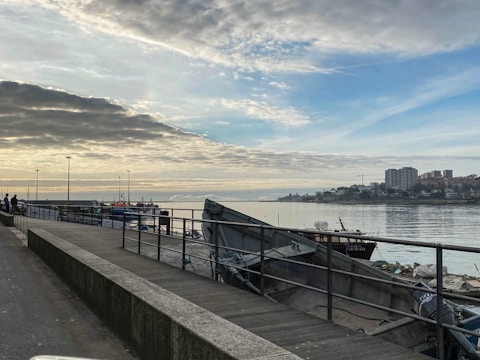 A waterfront scene featuring a paved walkway beside a body of water. A few people are visible on the left walking along the path. Several boats are docked along the pier, with masts and rigging visible against the sky. In the distance, there are buildings clustered together, possibly part of a small town or city. Above, the sky is partly cloudy, with patches of blue emerging between grey and white clouds.