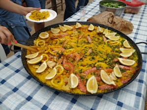 A large pan of paella on a checkered tablecloth with garnishes of lemon wedges, shrimp, calamari, and various vegetables. A person is serving a portion onto a white plate. Other food items and a glass are visible on the table.