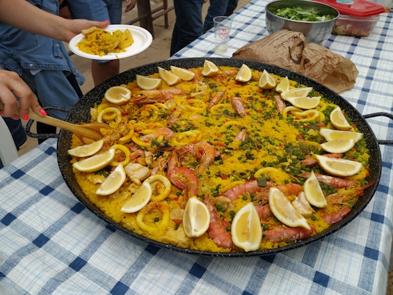 A large pan of paella on a checkered tablecloth with garnishes of lemon wedges, shrimp, calamari, and various vegetables. A person is serving a portion onto a white plate. Other food items and a glass are visible on the table.