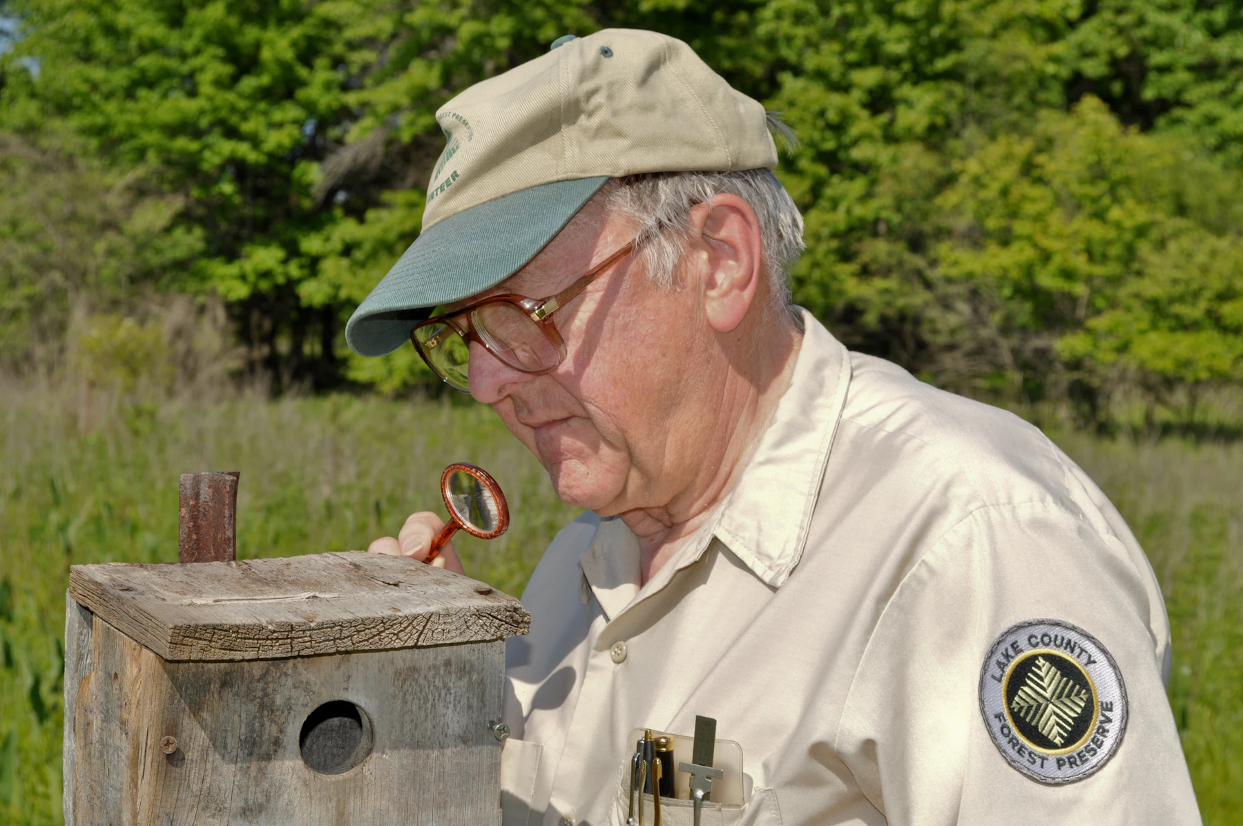 A man in a tan cap and uniform shirt bending over a weathered wooden nest box in a meadow, studying it closely through a small round glass in his hand