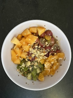 A bowl filled with a colorful mix of fruits including diced cantaloupe, red grapes, kiwi slices, orange segments, and topped with chopped nuts. The fruits are arranged in sections, providing a visually appealing contrast of colors and textures.