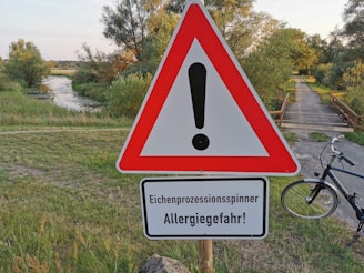 A warning sign with an exclamation mark in a red triangle is prominently displayed in a grassy area near a river or stream. Below the triangle, a sign in German warns about the Oak Processionary Moth and allergy risk. A pathway and a bicycle are visible to the right, with trees and greenery surrounding the area.