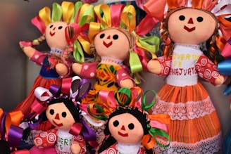 A collection of colorful handmade dolls with smiling faces and vibrant outfits. The dolls display various braided hairstyles with bright ribbon decorations. Each doll is uniquely dressed in traditional, intricately patterned clothing that adds charm and character to their appearance.