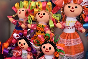 A collection of colorful handmade dolls with smiling faces and vibrant outfits. The dolls display various braided hairstyles with bright ribbon decorations. Each doll is uniquely dressed in traditional, intricately patterned clothing that adds charm and character to their appearance.
