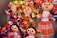 A collection of colorful handmade dolls with smiling faces and vibrant outfits. The dolls display various braided hairstyles with bright ribbon decorations. Each doll is uniquely dressed in traditional, intricately patterned clothing that adds charm and character to their appearance.